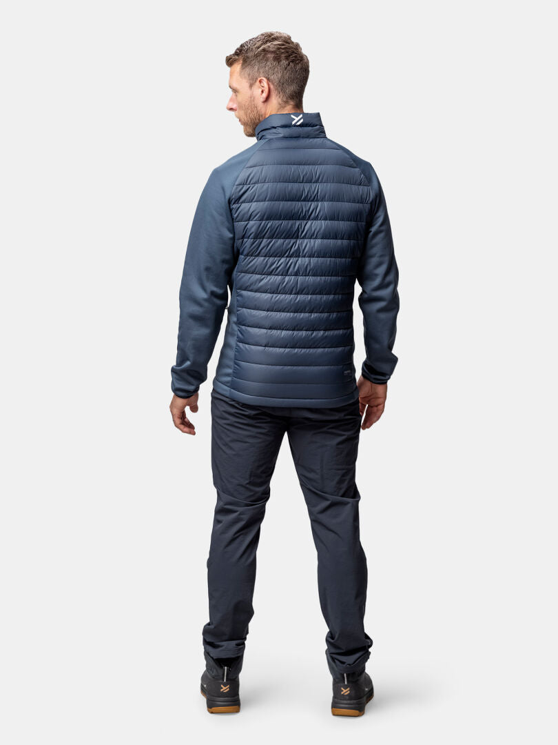 Ein Mann trägt die Halti Huippu Hybrid Daunenjacke Herren mit passender Hose, mit dem Gesicht zur Seite vor einem hellen Hintergrund.