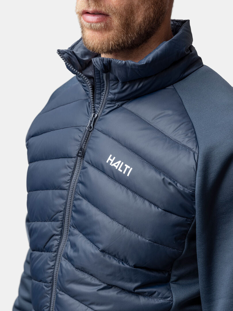 Ein Mann trägt eine Halti Huippu Hybrid Daunenjacke Herren in Marineblau, mit Reißverschluss vorne und hohem Kragen, von den Schultern aufwärts gezeigt.