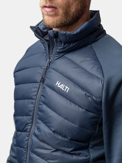 Ein Mann trägt eine Halti Huippu Hybrid Daunenjacke Herren in Marineblau, mit Reißverschluss vorne und hohem Kragen, von den Schultern aufwärts gezeigt.