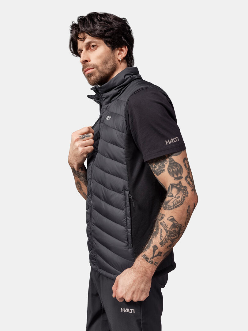 Mann mit Tattoos in schwarzer Halti Huippu Daunenweste, Pertex Quantum Ripstop, und T-Shirt, seitlich stehend.