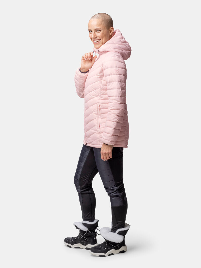 Frau in Halti Huippu Mid Daunenjacke, schwarzen Hosen und Winterstiefeln lächelt vor neutralem Hintergrund.