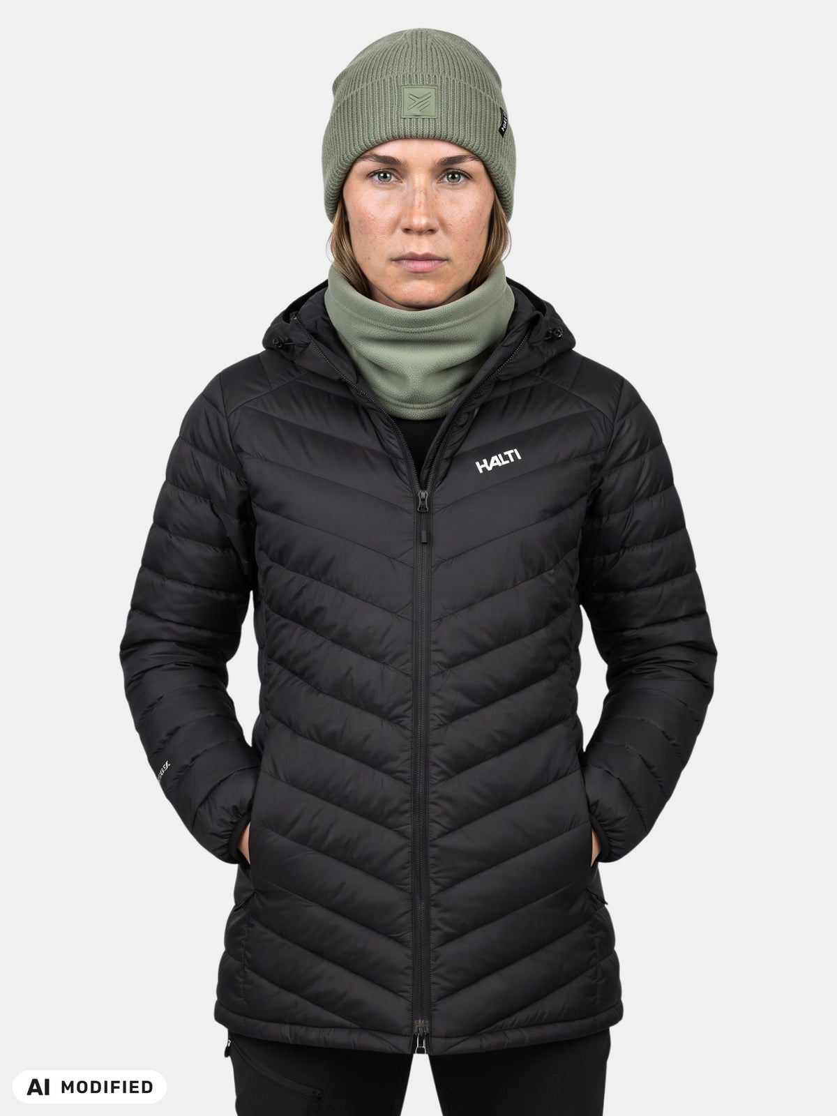 Eine Frau in einer schwarzen Halti Huippu Mid Daunenjacke, einer grünen Mütze und einem Halswärmer steht mit dem Gesicht nach vorne.