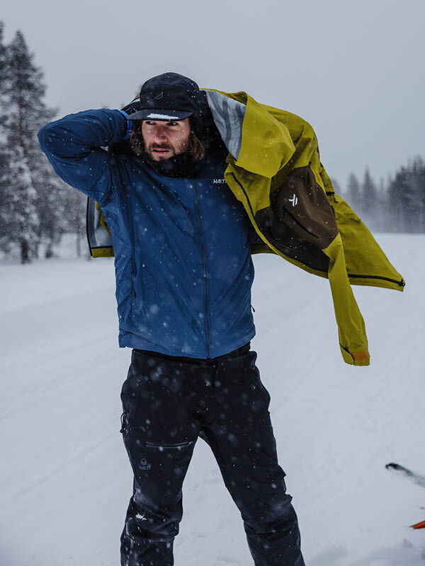 Ein Mann zieht seine gelbe Halti Haave Insulation Primaloft Jacke Herren über einer blauen Jacke inmitten verschneiter, winterlicher Bäume zurecht.
