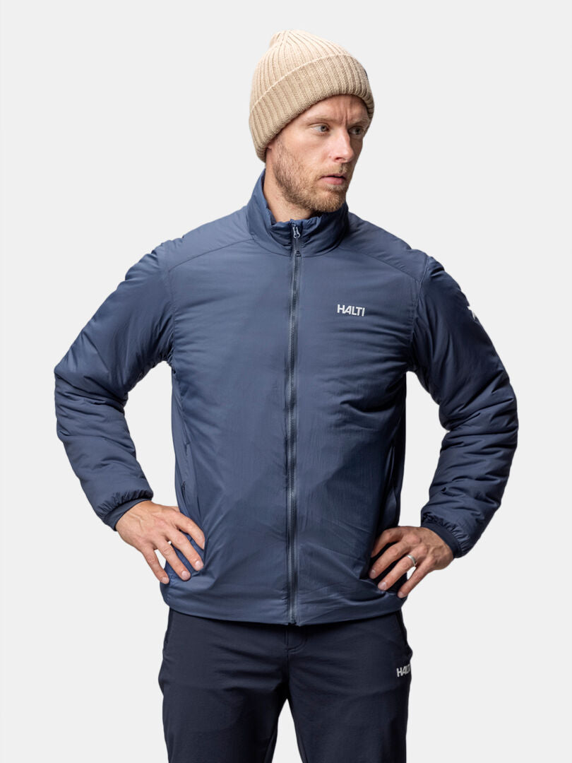 Ein Mann in einer blauen Halti Haave Insulation Primaloft Jacke Herren steht mit den Händen in den Hüften und blickt zur Seite.