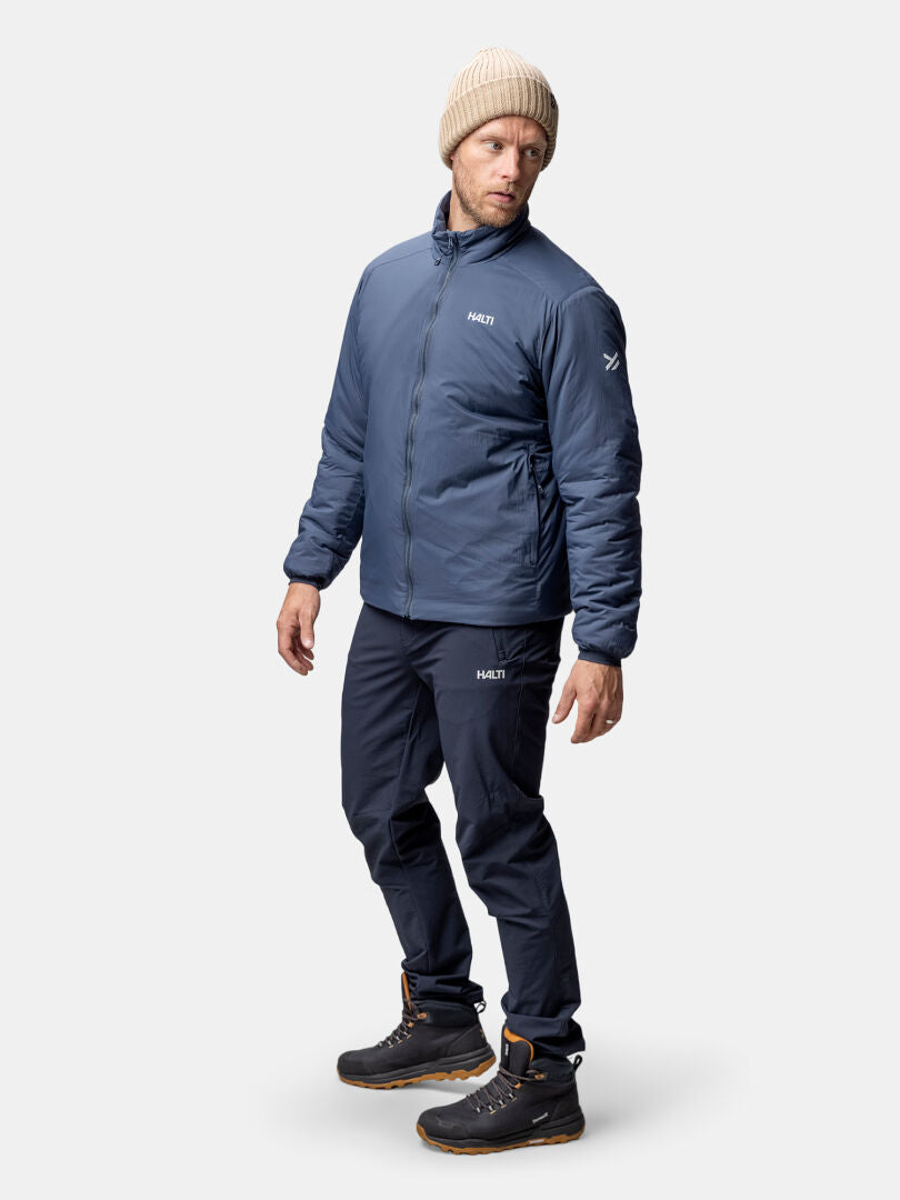 Ein Mann in einer blauen Halti Haave Insulation Primaloft Jacke Herren, dunkler Hose und Stiefeln steht vor einem schlichten Hintergrund.