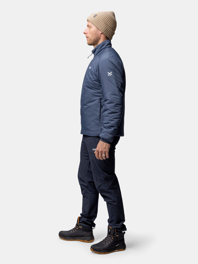 Ein Mann in einer Halti Haave Insulation Primaloft Jacke, einer marineblauen Hose und Wanderschuhen steht im Profil vor einem schlichten Hintergrund.