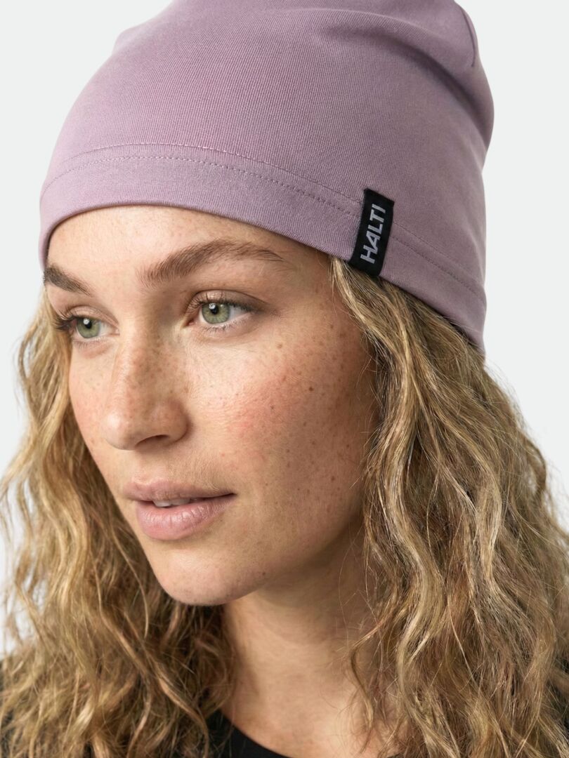 Eine Person mit gewelltem blondem Haar trägt eine hellviolette Halti Loiva Mütze und schaut leicht zur Seite.