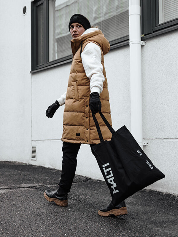 Person in hellbrauner Pufferweste, weißem Kapuzenpulli, schwarzer Mütze und Halti Viiri Fleece Handschuhen geht mit einer großen Tragetasche.