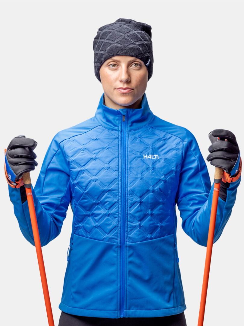 Eine Person in einer blauen Jacke, schwarzen Handschuhen und Halti Crostie Merino Mütze hält orangefarbene Skistöcke vor einem unifarbenen Hintergrund.