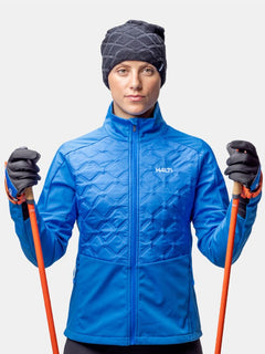 Eine Person in einer blauen Jacke, schwarzen Handschuhen und Halti Crostie Merino Mütze hält orangefarbene Skistöcke vor einem unifarbenen Hintergrund.