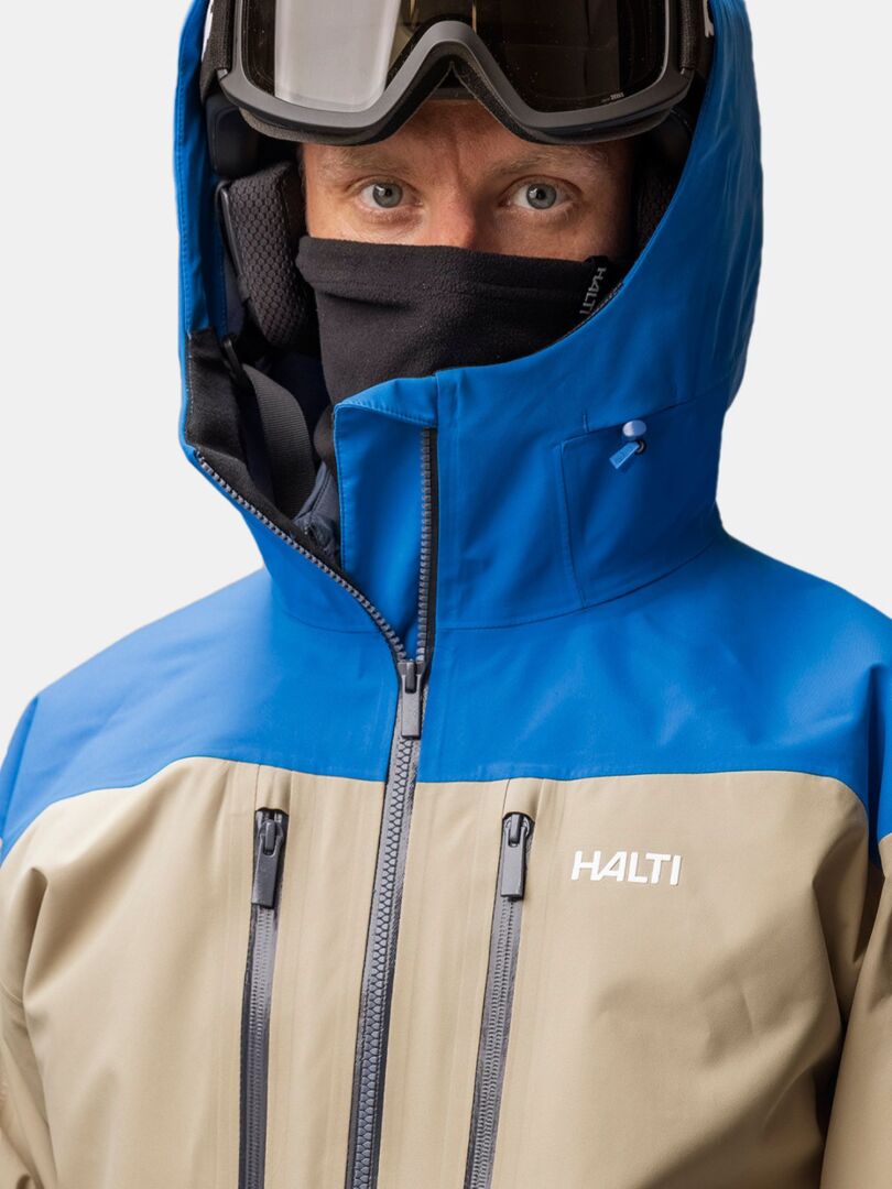 Eine Person, die einen Halti Trone Fleece Schlauchschal unter einer blau-beigen Skijacke mit Kapuze und einer Skibrille trägt.