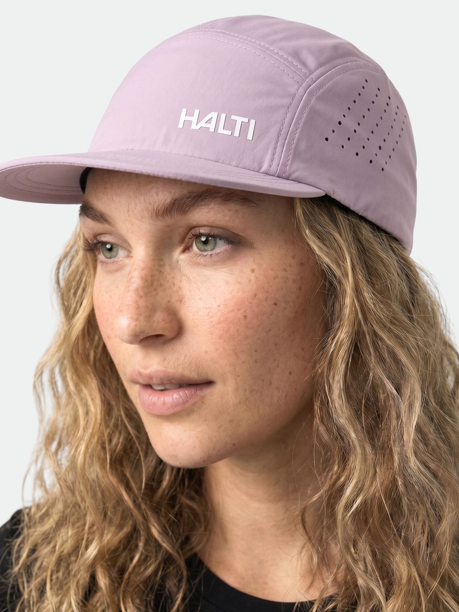 Eine Frau mit lockigem blondem Haar trägt eine hellviolette Halti Luovi Cap und schaut leicht zur Seite.