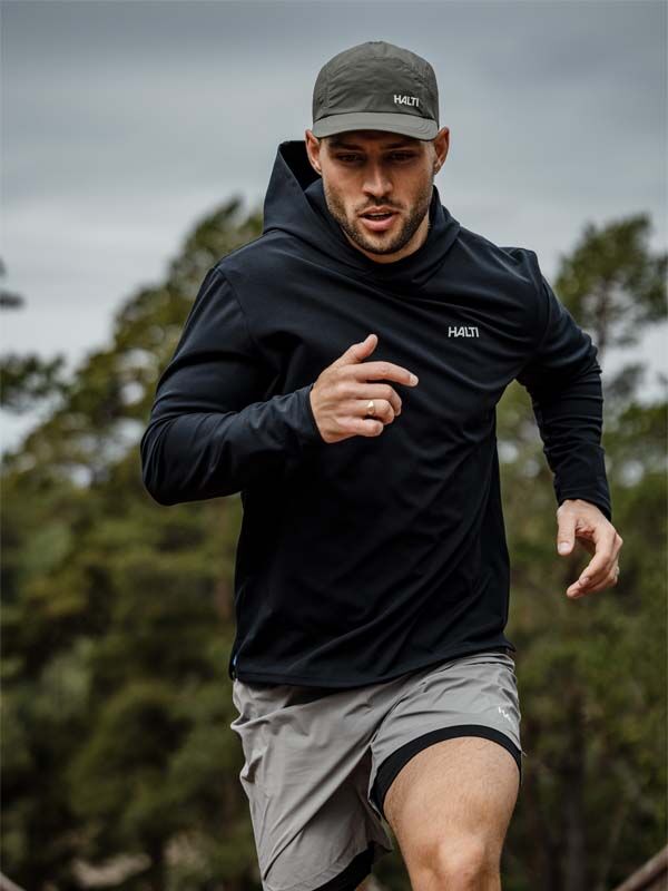 Ein Mann läuft im Freien in einem schwarzen Kapuzenpulli, grauen Shorts und einer Halti Luovi Cap, mit Bäumen im Hintergrund.