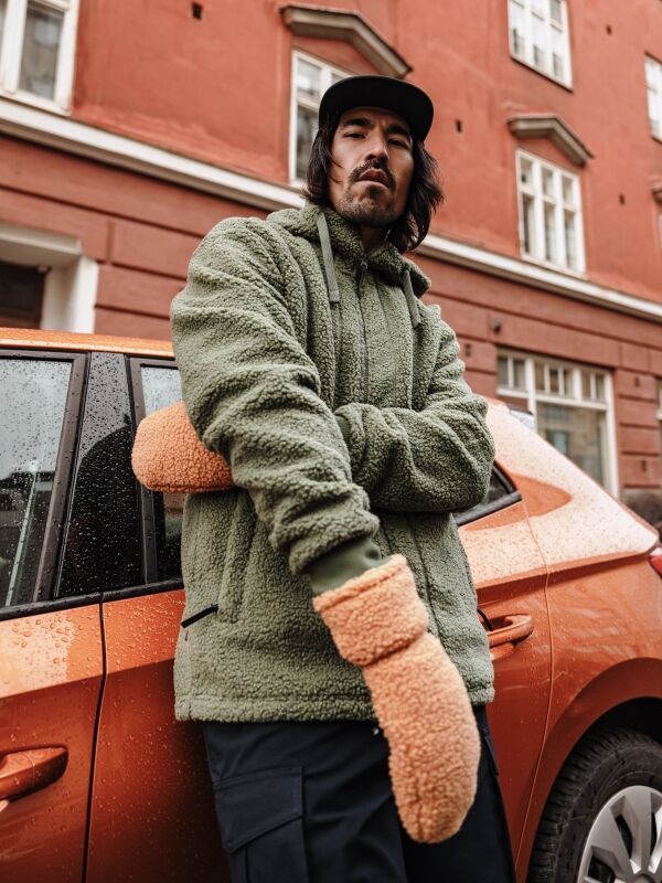 Ein Mann in grüner Fleecejacke und Halti Lykky Fleece Fäustlinge steht bei einem orangefarbenen Auto in der Nähe eines roten Gebäudes.
