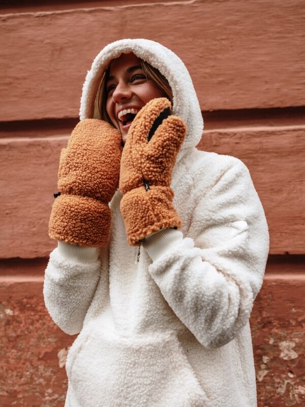 Person in weißem Sherpa-Kapuzenpulli und Halti Lykky Fleece Fäustlinge lächelt vor einer strukturierten rötlich-braunen Wand.