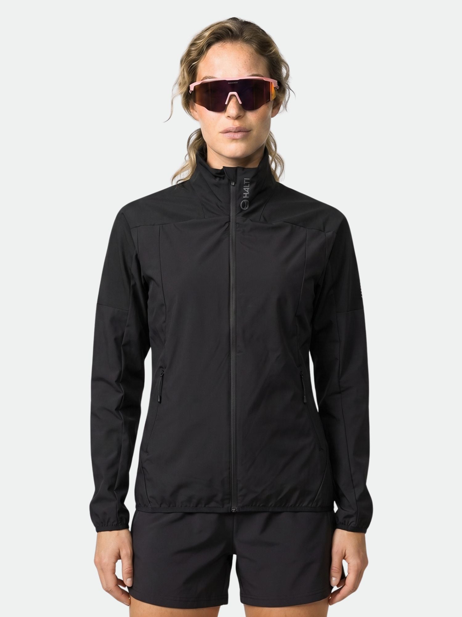 Eine Frau trägt die Halti Urbanite Damen Training Jacke mit schwarzen Shorts und Sonnenbrille vor einem hellen Hintergrund.