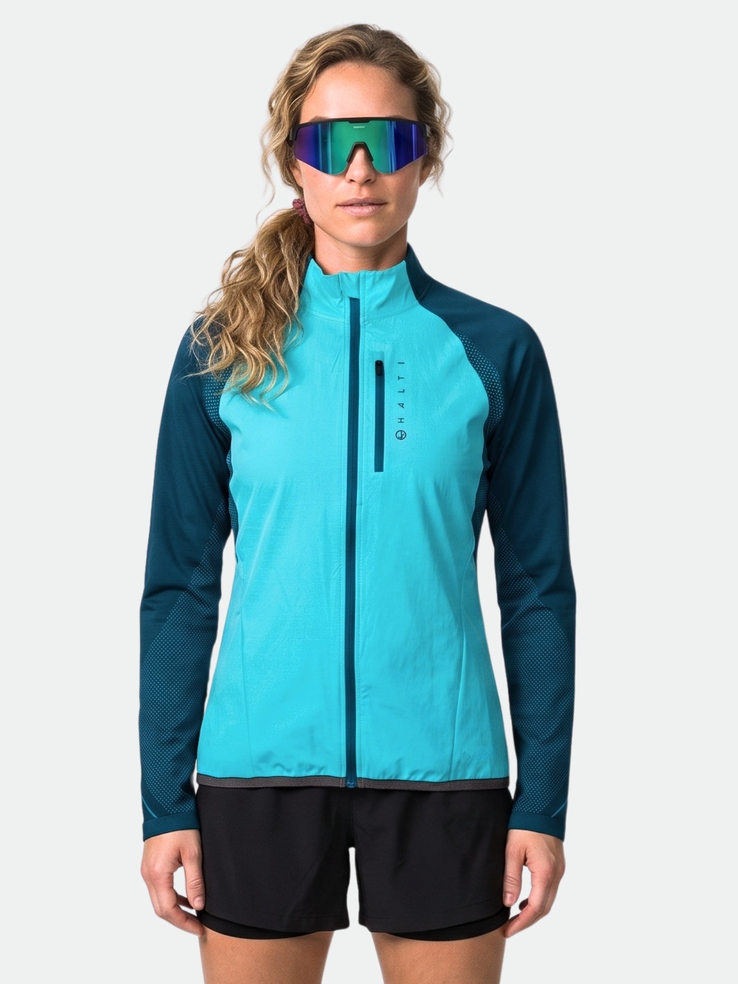 Eine Frau trägt vor einem einfarbigen Hintergrund die „Halti Tracks Damen Seamless Jacke“, schwarze Shorts und eine reflektierende Sonnenbrille.