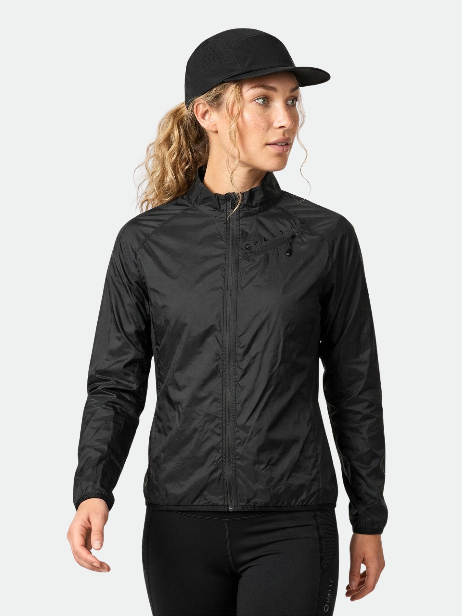 Frau trägt eine Halti Control Damen Windbreaker Jacke in schwarz mit Leggings, stehend vor einem hellgrauen Hintergrund.