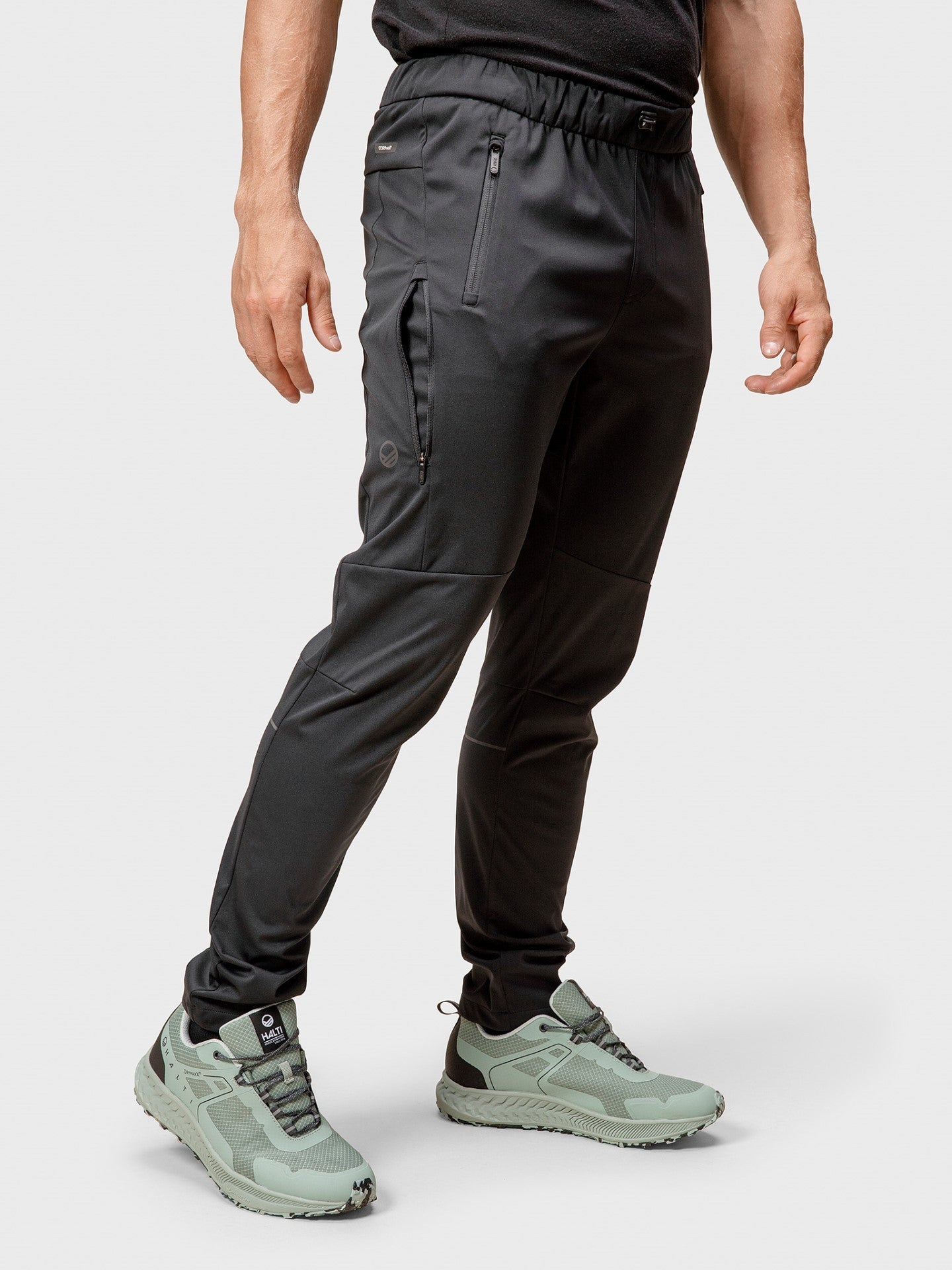 Ein Mann in Halti Exhale Stormwall Hosen und Hemd, mit hellgrünen Sportschuhen, steht vor einem schlichten Hintergrund.