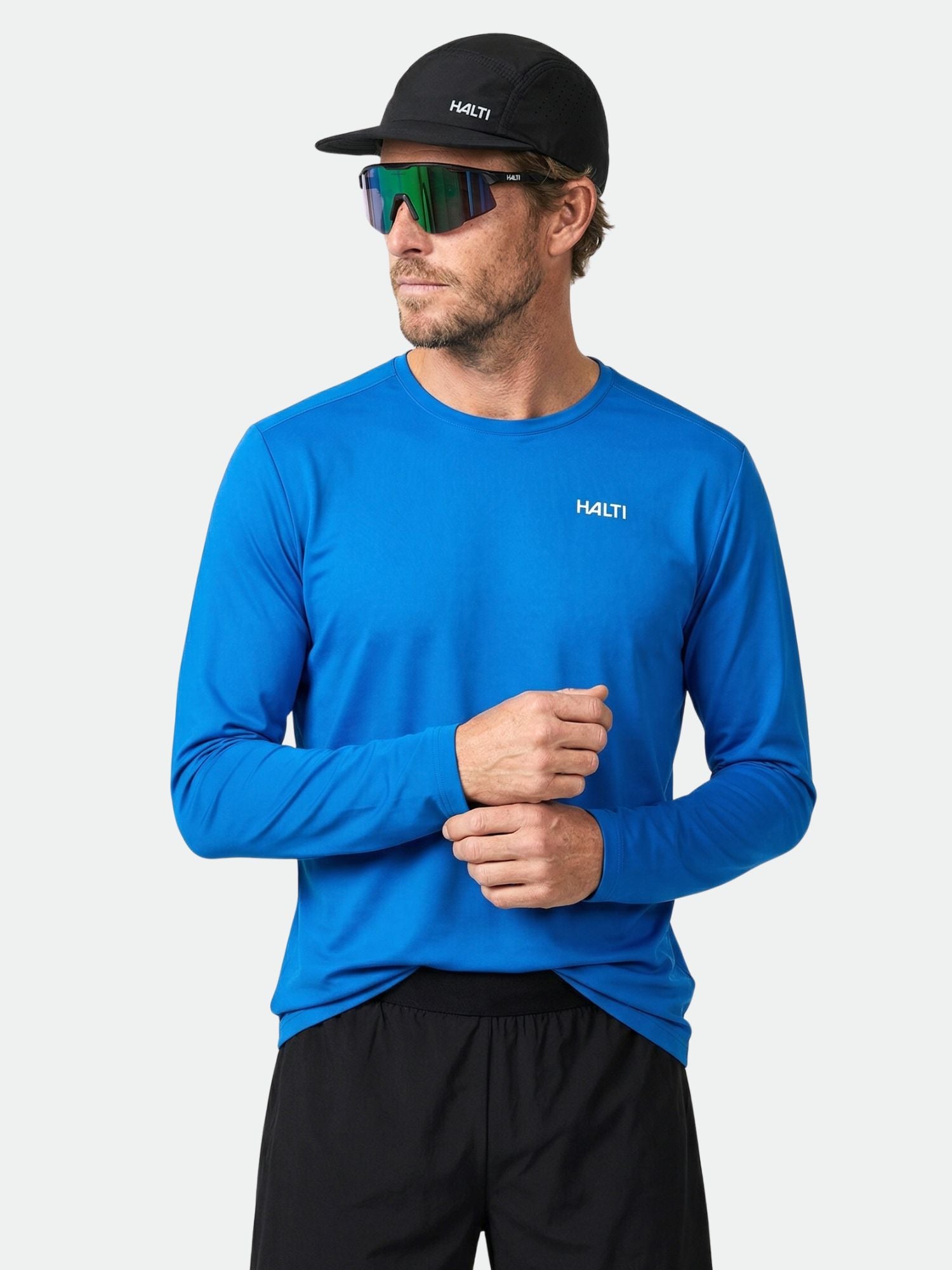 Ein Mann in einem Halti Salves Trainingsshirt Herren, schwarzem Hut und Sonnenbrille steht vor einem schlichten hellen Hintergrund.