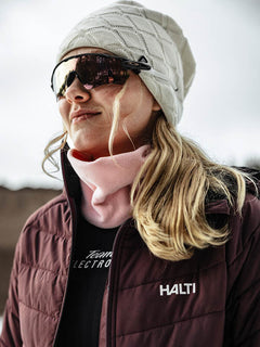 Frau mit Sonnenbrille, Halti Crostie Merino Mütze, rosa Halswärmer und kastanienbrauner Halti Jacke im Freien.