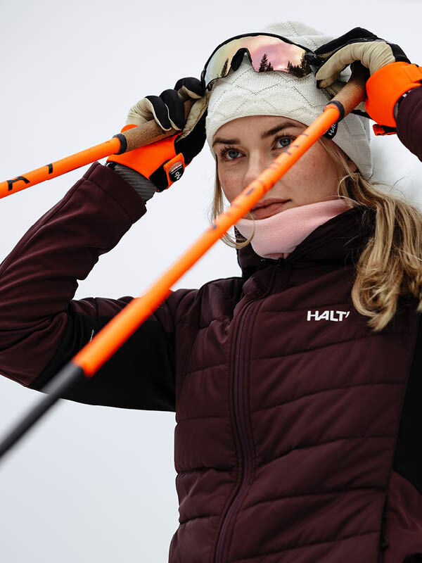 Eine Frau mit Halti Crostie Merino Mütze rückt ihre Skibrille zurecht und hält orangefarbene Skistöcke vor einem schlichten Hintergrund.