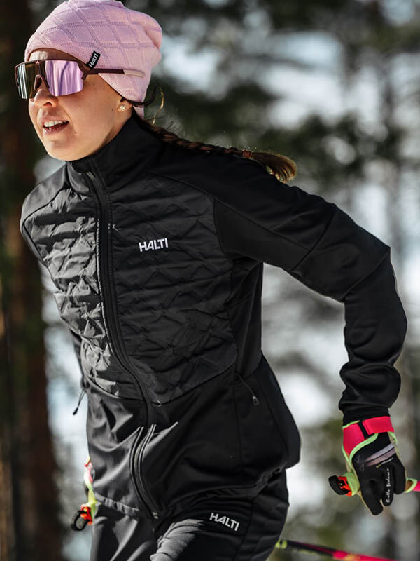 Eine Person in einer schwarzen Halti Framover Hybrid Langlaufjacke Damen, einem rosa Hut und einer Sonnenbrille ist beim Skilanglauf im Freien.