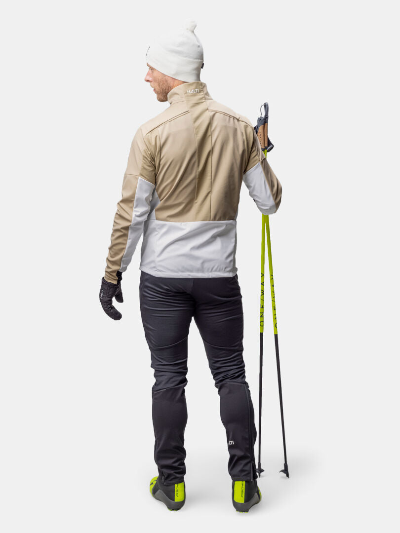 Mit der Halti Framover Hybrid Langlaufjacke Herren ist ein Mann mit Skistöcken bereit zum Skilanglauf vor einem schlichten Hintergrund.