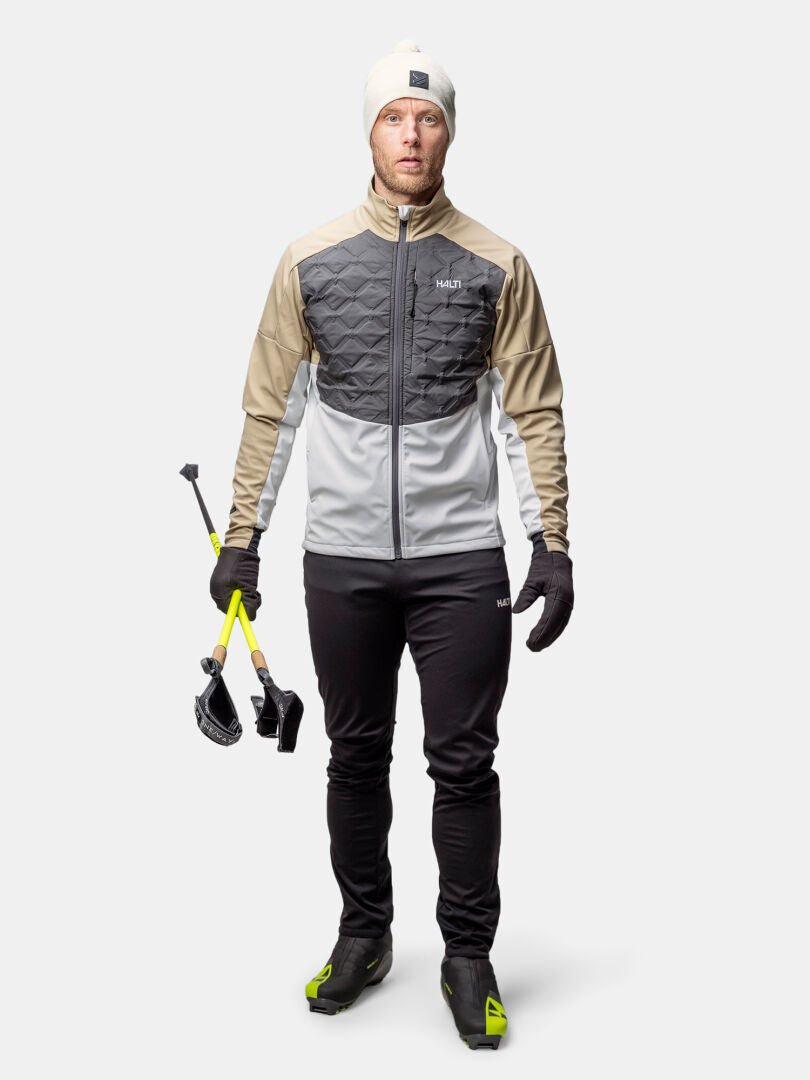 Mann in Halti Framover Hybrid Langlaufjacke mit Skistöcken, bereit zum Skilanglauf vor grauem Hintergrund.