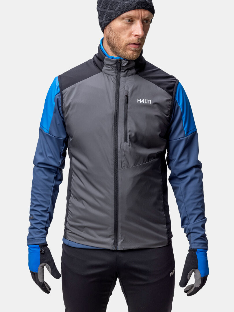 Ein Mann in einer grau-blauen Halti Framover Langlaufweste Unisex, schwarzen Handschuhen und Mütze steht mit dem Gesicht nach vorne.