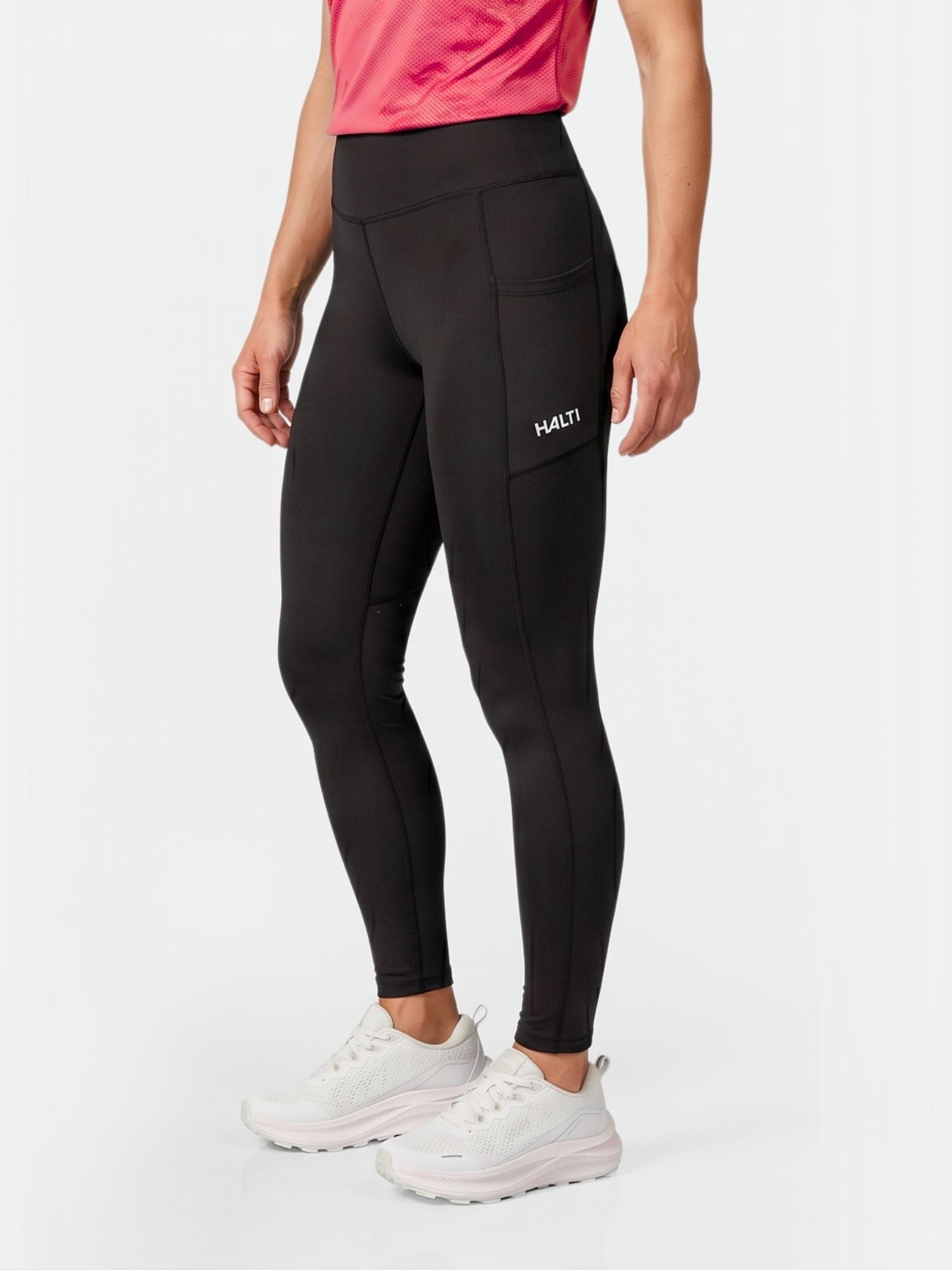 Eine Frau in Halti Kaldu Training Tights Damen mit Seitentaschen, einem rosa Hemd und weißen Turnschuhen steht vor einem schlichten Hintergrund.