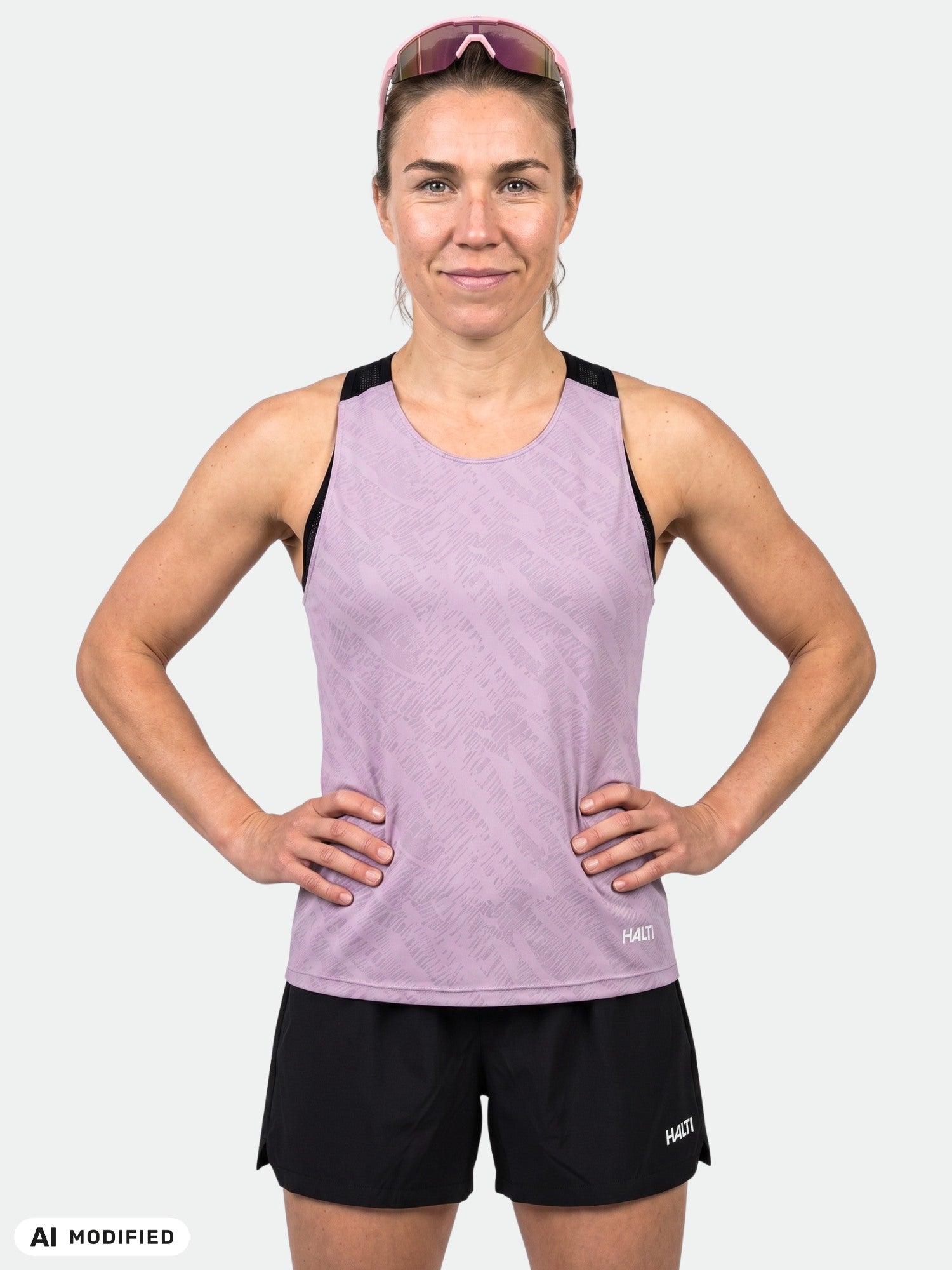 Eine lächelnde Frau in einem „Halti Oulanka Trainingstop Damen“ steht mit den Händen in die Hüften gestemmt vor einem einfarbigen Hintergrund.