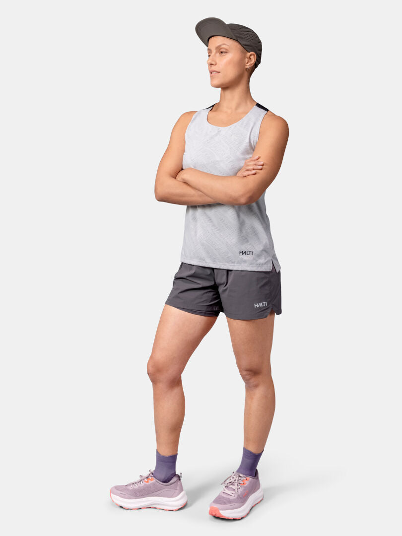 Eine Frau in einem grauen Halti Oulanka Trainingstop Damen, dunklen Shorts, Mütze und lila Turnschuhen steht mit verschränkten Armen.