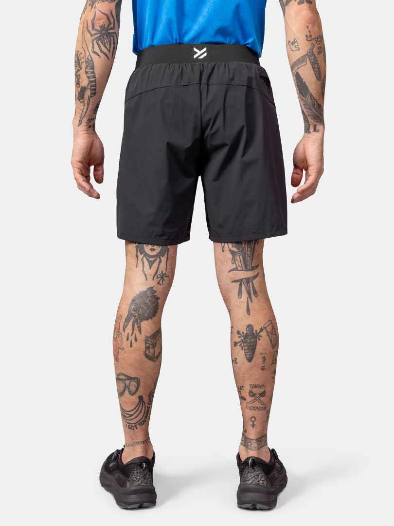 Eine Person in Halti Uurto Shorts Herren und schwarzen Schuhen wird von hinten gezeigt, wobei tätowierte Arme und Beine zu sehen sind.