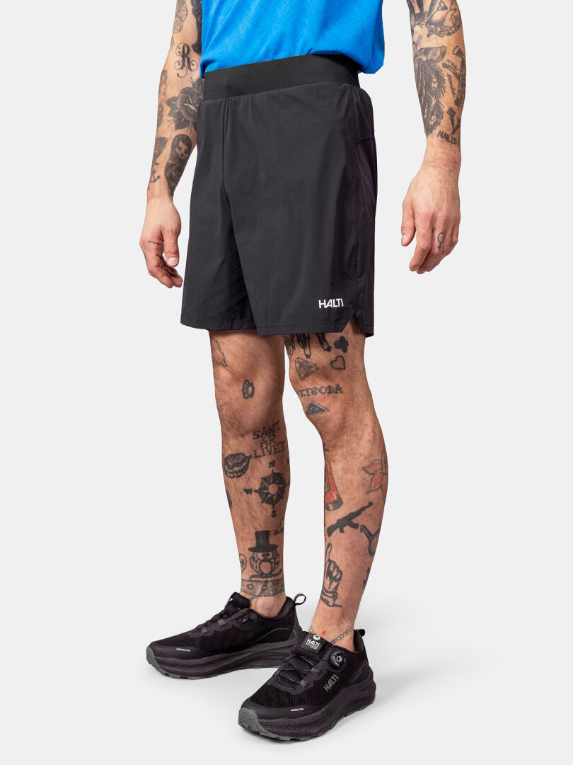 Eine Person in blauem Hemd und Halti Uurto Shorts Herren, schwarze Laufschuhe, Tattoos an Armen und Beinen.