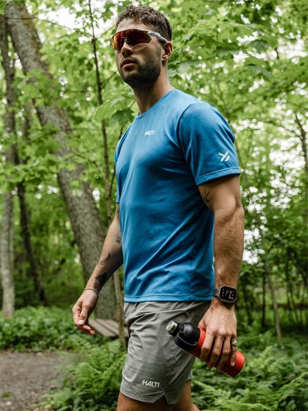 Mann trägt Halti Uurto Shorts Herren beim Walken im Wald, mit Wasserflasche, Sonnenbrille und Smartwatch.