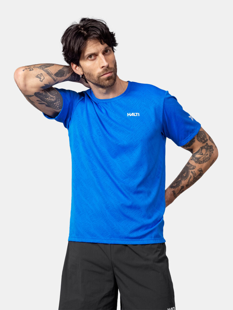 Ein Mann in einem blauen Halti Oulanka T-Shirt und schwarzen Shorts steht mit einer Hand im Nacken vor einem schlichten Hintergrund.