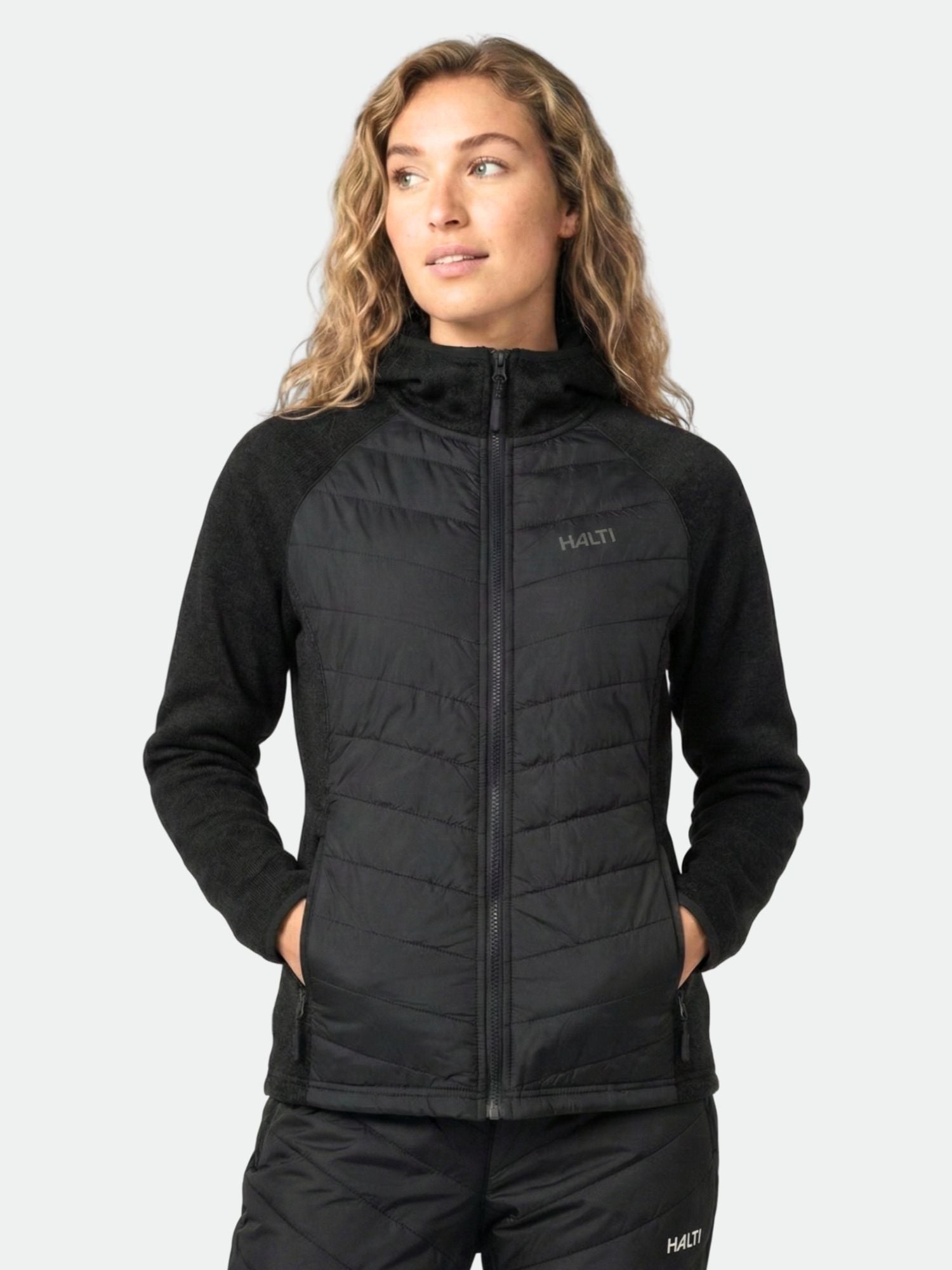 Eine Frau in der Halti Streams Hybrid Midlayer Jacke Damen mit den Händen in den Taschen, mit Blick zur Seite - ideal für Outdoor-Aktivitäten.