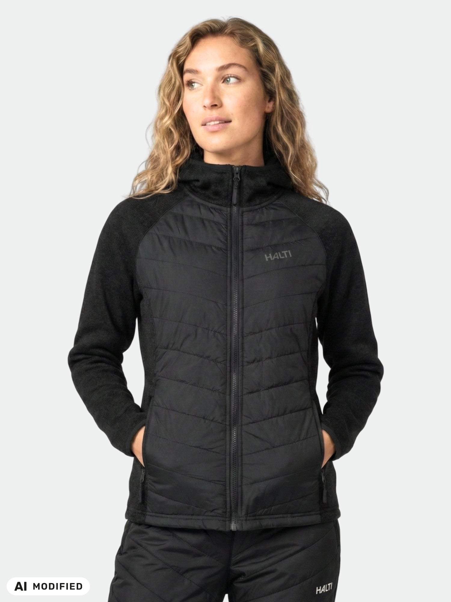 Eine Frau in einer schwarzen Halti Streams Hybrid Midlayer Jacke für Damen steht mit den Händen in den Taschen vor einem hellgrauen Hintergrund.