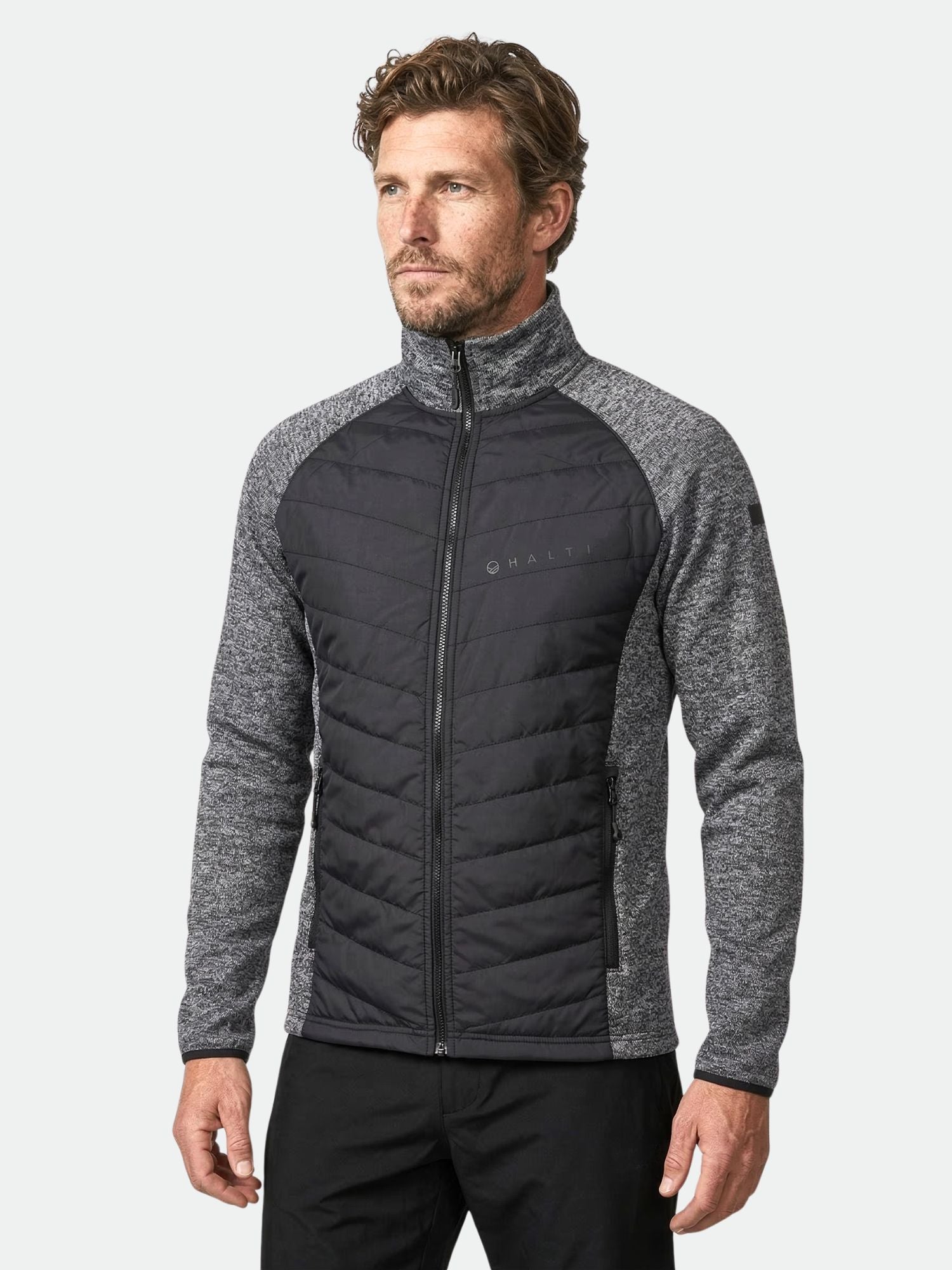 Ein Mann trägt die Halti Streams Hybrid Layer Jacke Herren in Grau und Schwarz mit hohem Kragen, vor einem unifarbenen Hintergrund.