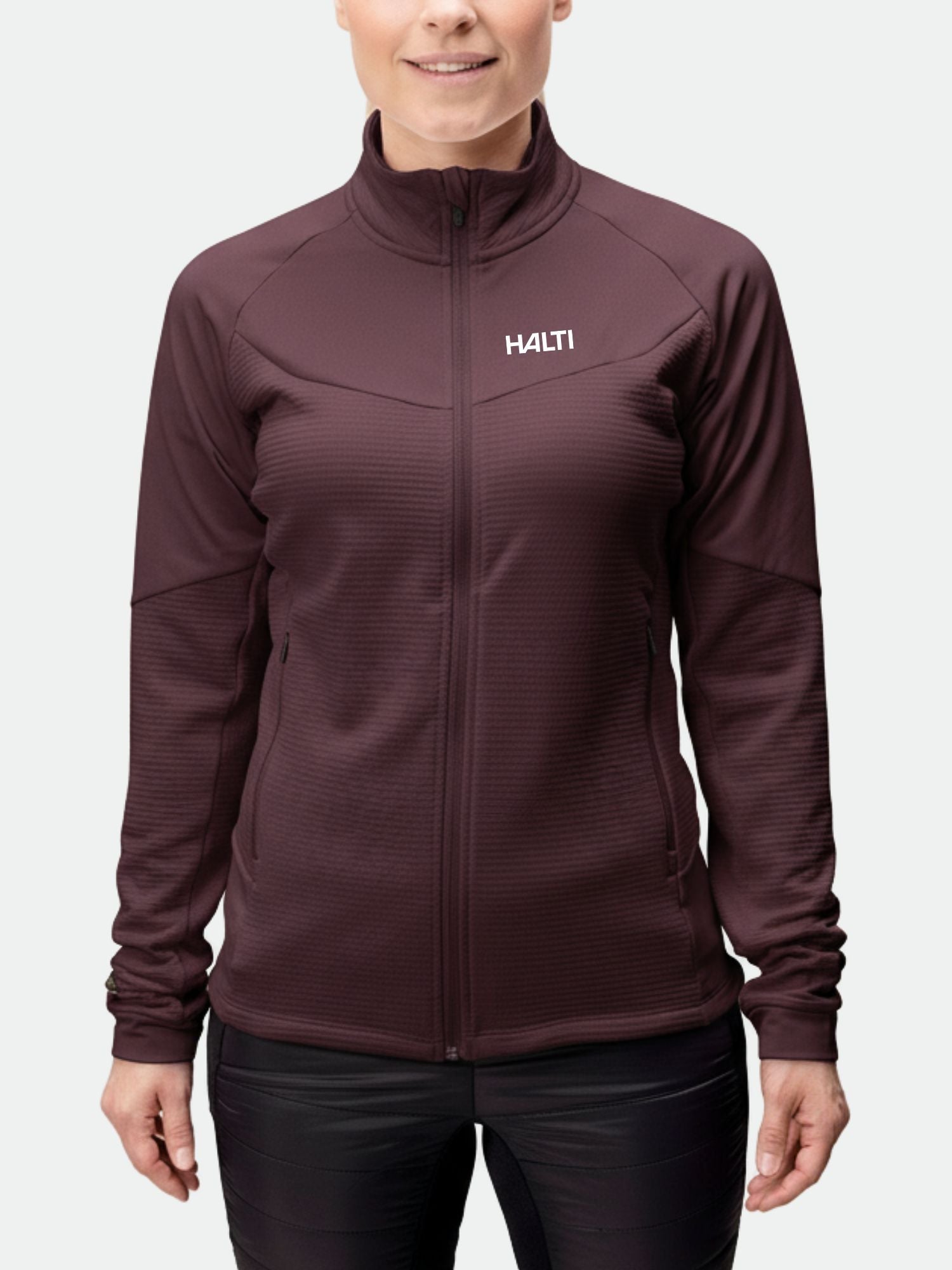 Eine Frau trägt eine kastanienbraune Halti HeatGrid Midlayer Jacke Damen, ideal für Wanderungen, im Stehen und mit entspannten Händen.