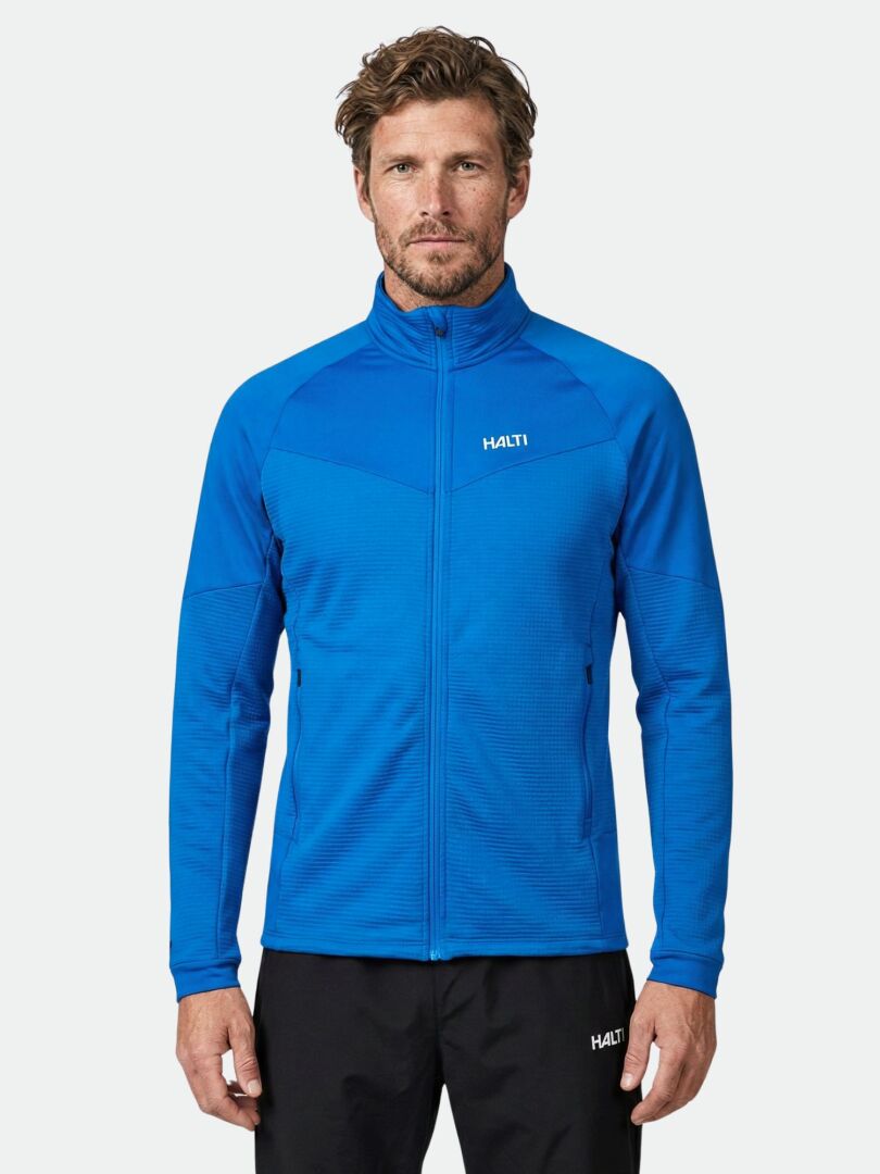 Ein Mann trägt eine blaue Halti HeatGrid Midlayer Jacke Herren und eine schwarze Hose und steht vor einem schlichten hellen Hintergrund.