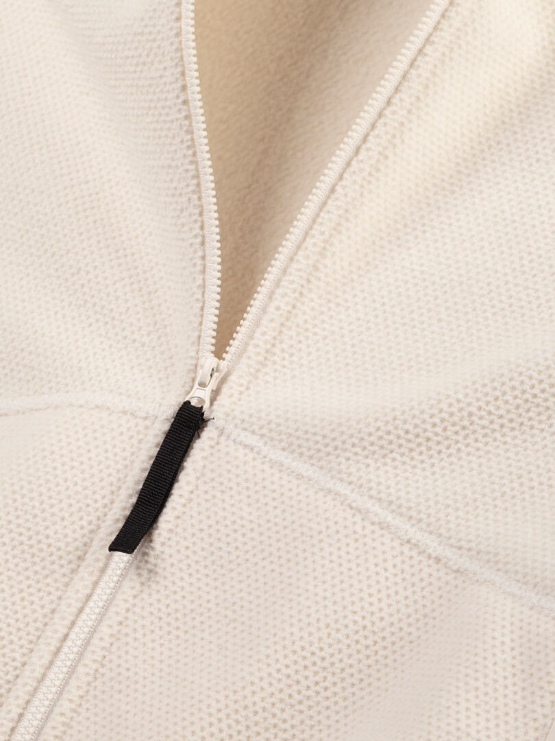 Nahaufnahme der Halti Soolo Midlayer Jacke Damen, teilweise geöffnet, zeigt cremefarbenes strukturiertes Gewebe und schwarzen Reißverschluss.