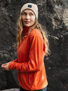 Eine Person in orangefarbener Jacke und Halti Vataja Mütze steht im Freien vor Felsen und schaut zur Seite.