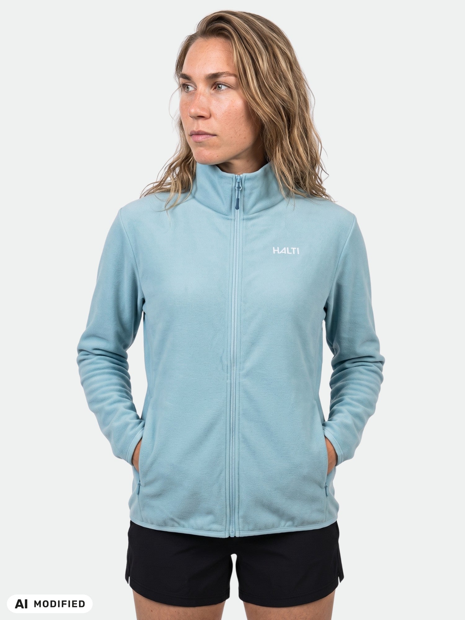 Frau in einer „Halti Rockmoon Fleecejacke Damen“ und schwarzen Shorts, die Hände in den Taschen, vor einem einfarbigen Hintergrund.