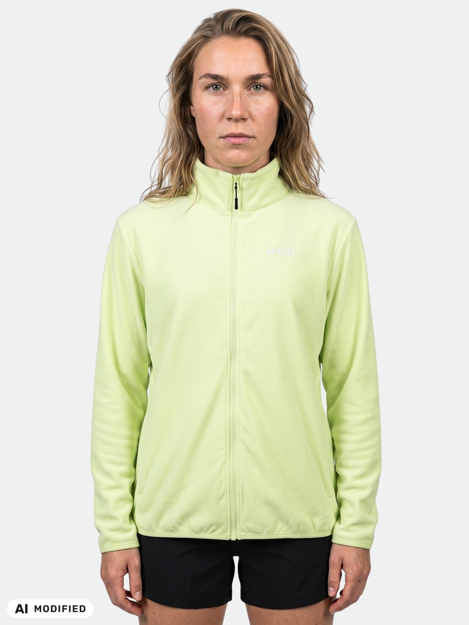 Eine Person trägt eine hellgrüne Halti Rockmoon Fleecejacke für Damen und schwarze Shorts und blickt vor einem einfarbigen Hintergrund nach vorne.