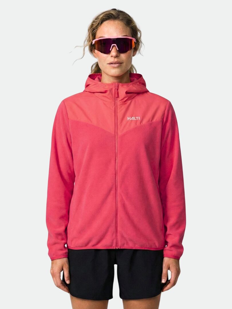 Eine Frau trägt die Halti Rockmoon Fleecejacke Damen in Pink, schwarze Shorts und eine Sonnenbrille vor einem schlichten Hintergrund.