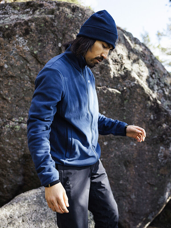 Eine Person in einer blauen Halti Rockmoon Fleecejacke Herren, einer schwarzen Hose und einer blauen Mütze steht bei großen Felsen im Freien.