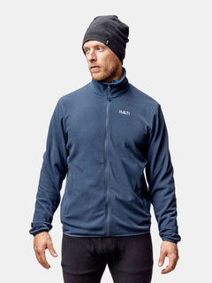 Ein Mann mit einer grauen Mütze, einer schwarzen Hose und einer Halti Rockmoon Fleece Jacke Herren steht vor einem hellen Hintergrund.