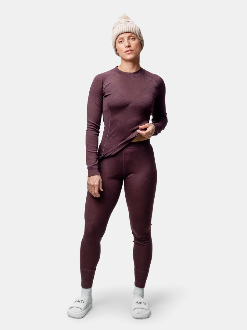 Eine Person trägt das Halti Hossa II Merino Unterwäsche Set Damen, eine beige Strickmütze und weiße Hausschuhe auf einem unifarbenen Hintergrund.